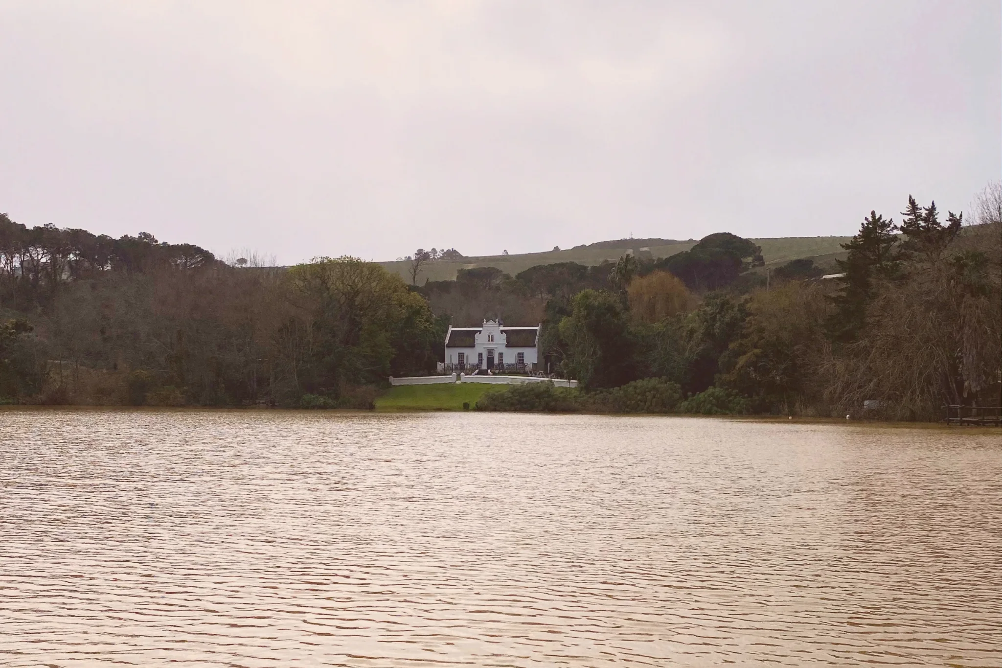 Zevenwacht’s white Cape Dutch manor house is seen across a lake, surrounded by lawns, mature trees and rolling green hills.