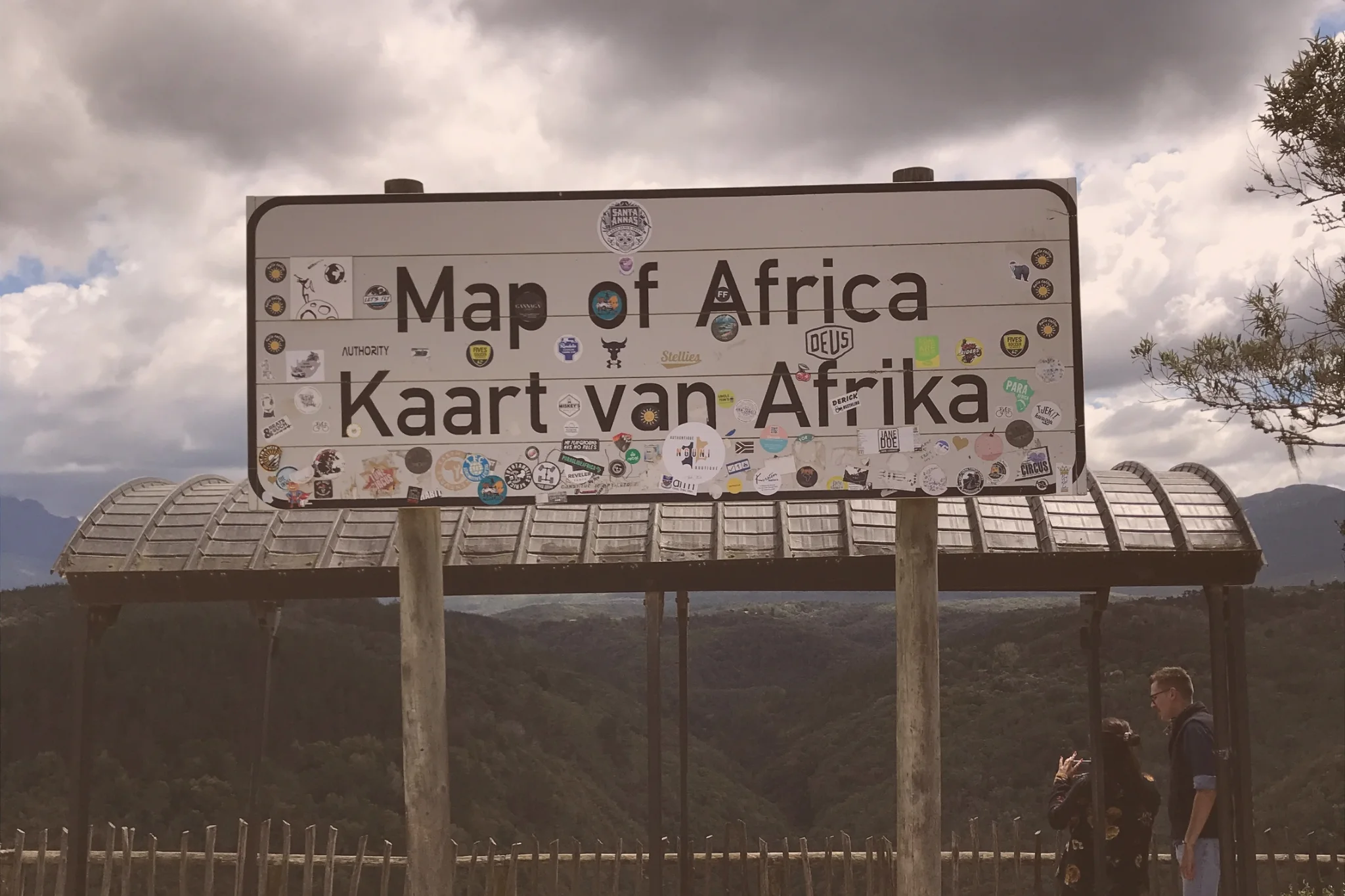 Two visitors stand beside the sticker-covered Map of Africa viewpoint sign in Wilderness, with a wooded valley and cloudy skies behind them.