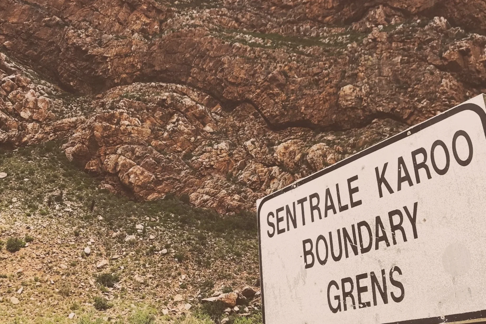 A roadside sign reading “Sentrale Karoo Boundary Grens” stands beneath a steep mountain face with visibly folded rock layers.