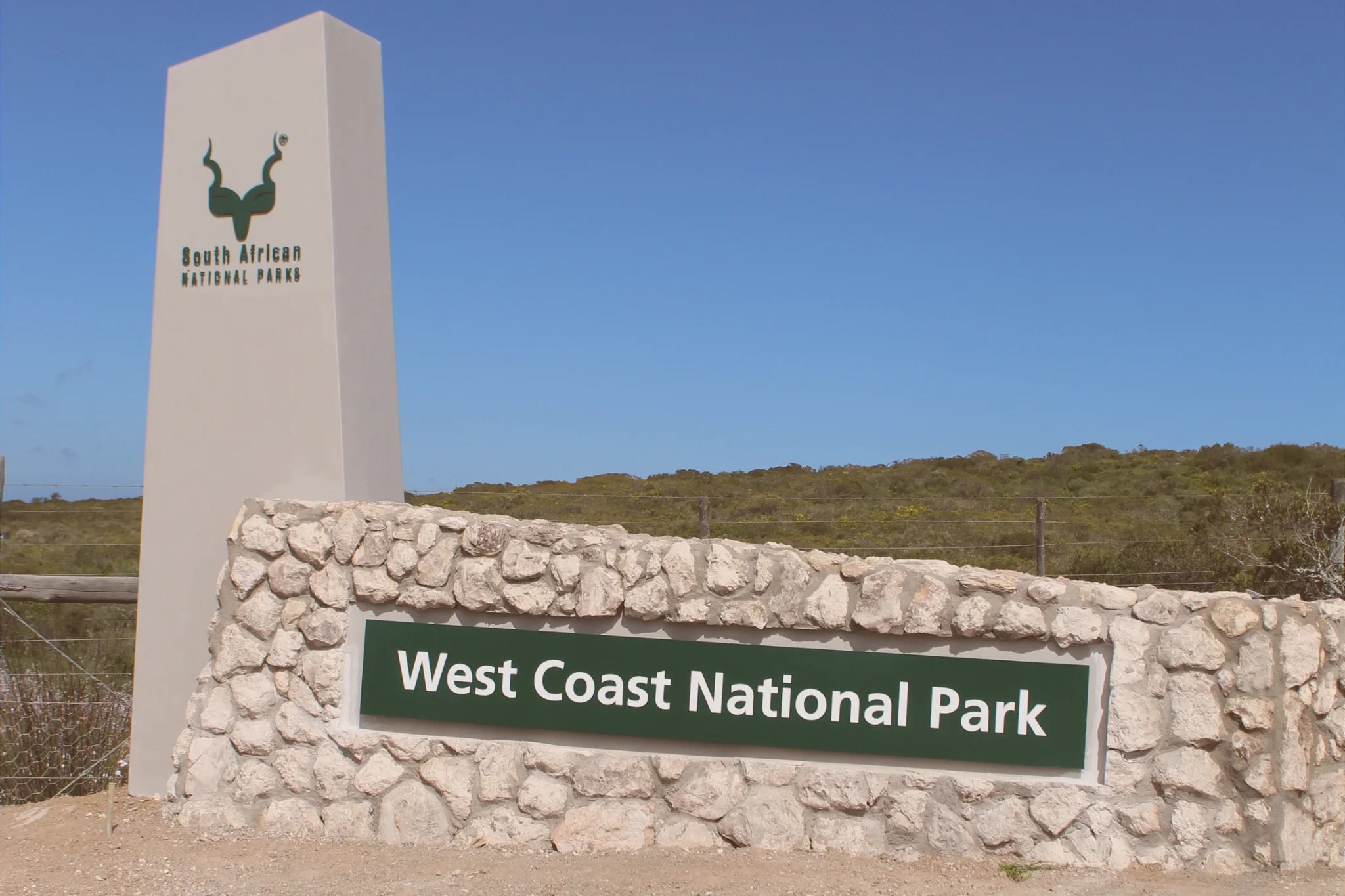West Coast National Park entrance sign set into a stone wall beside a tall South African National Parks pillar beneath a clear blue sky.