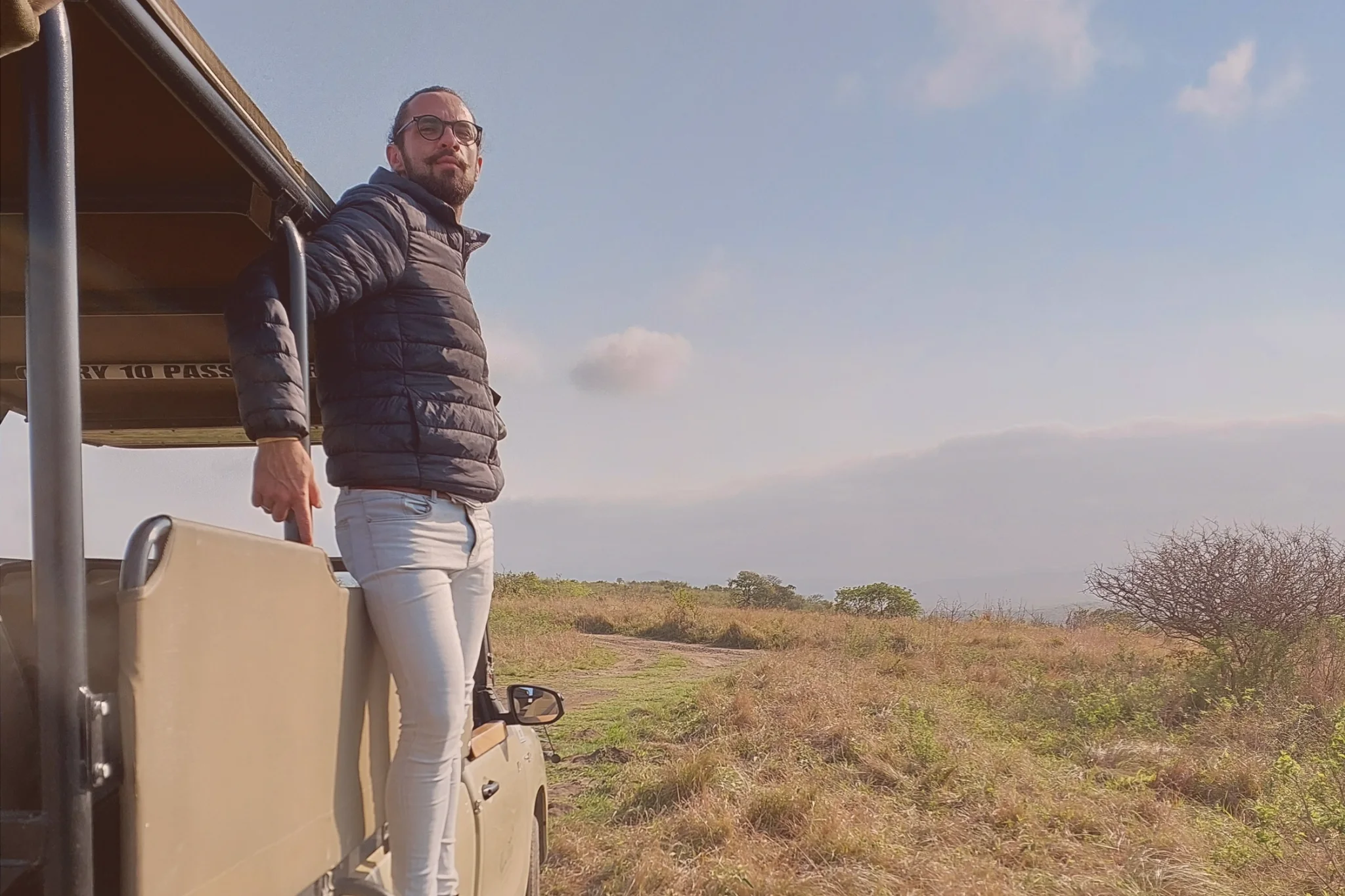 A traveller wearing glasses and a dark padded jacket stands in an open safari vehicle overlooking sunlit grassland in Hluhluwe, South Africa.