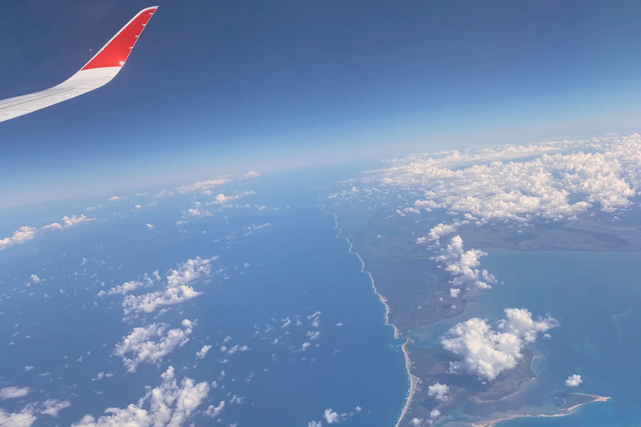 Air Mauritius red-tipped aircraft wing above scattered clouds, a sandy coastline and turquoise coastal waters, photographed on a flight to Mauritius.
