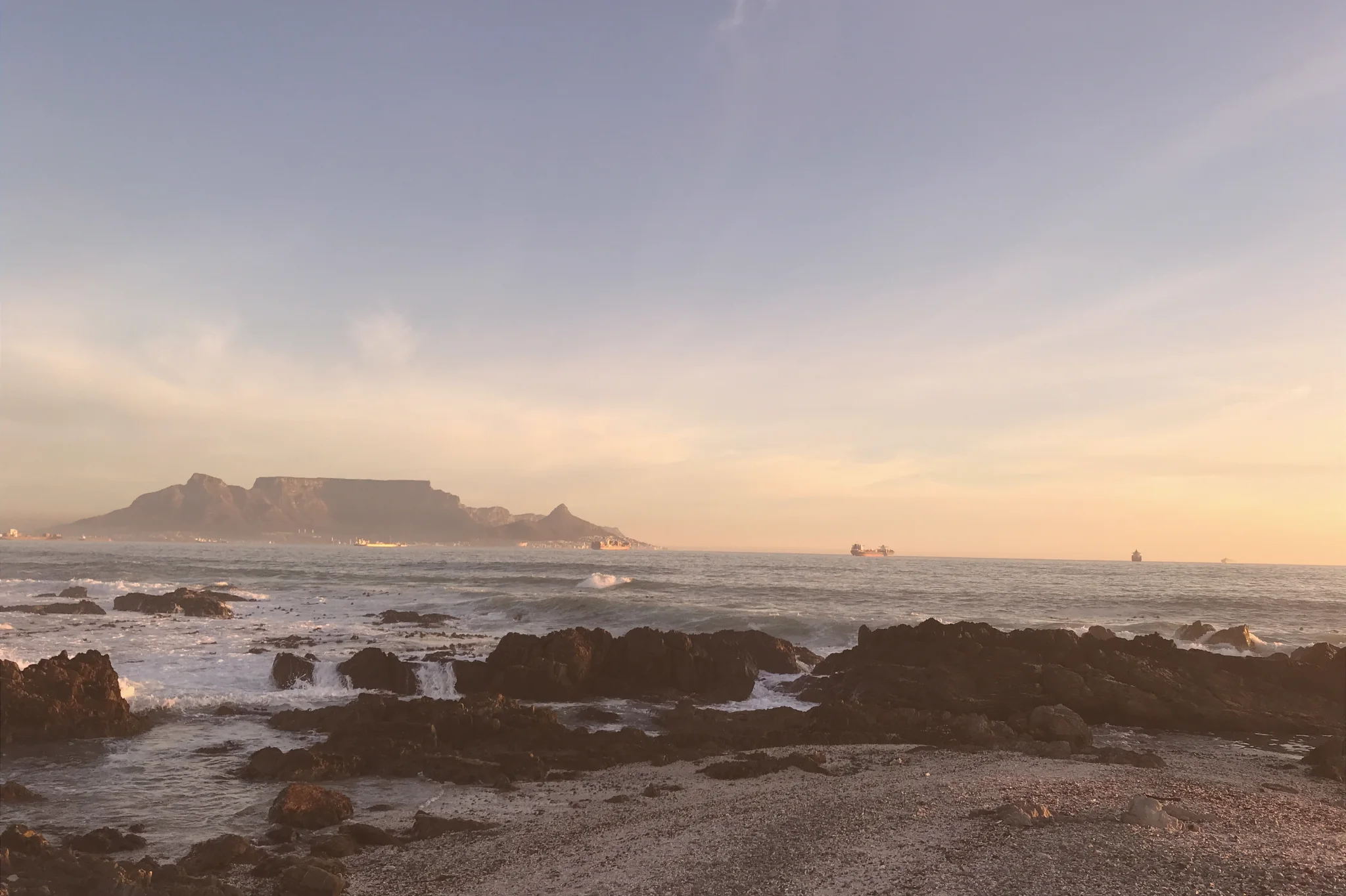 Table Mountain and Lion’s Head rise across Table Bay, with waves breaking over dark foreground rocks beneath a softly lit sky.